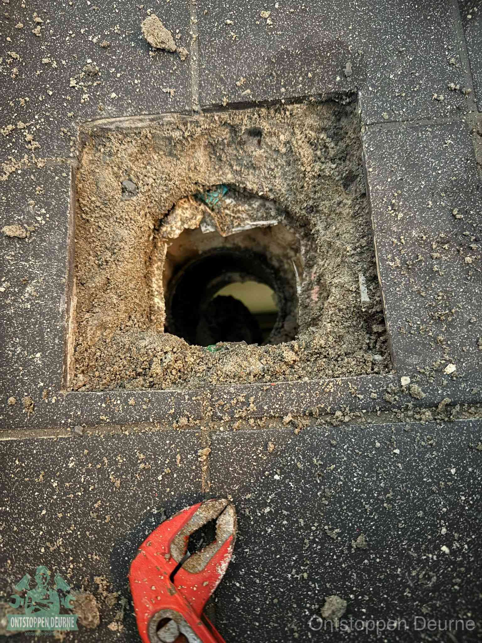 Een geopend rioolputje in een betontegelvloer met losgemaakt rooster en een zichtbare ronde afvoerbuis, het achterhalen waar de afvoerverstopping zit in Deurne
