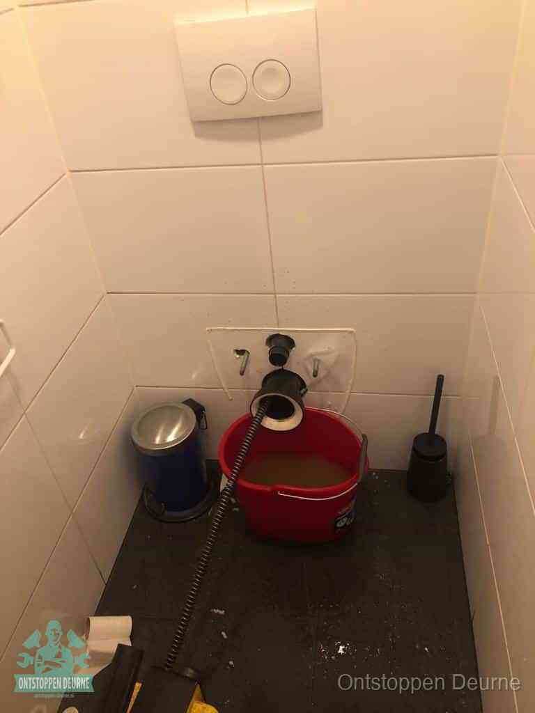 Een ontstopte wand-wc met verwijderde toiletpot waar een ontstoppingsveer in de wanduitgang verdwijnt, het achterhalen waar de verstopping zit in Deurne