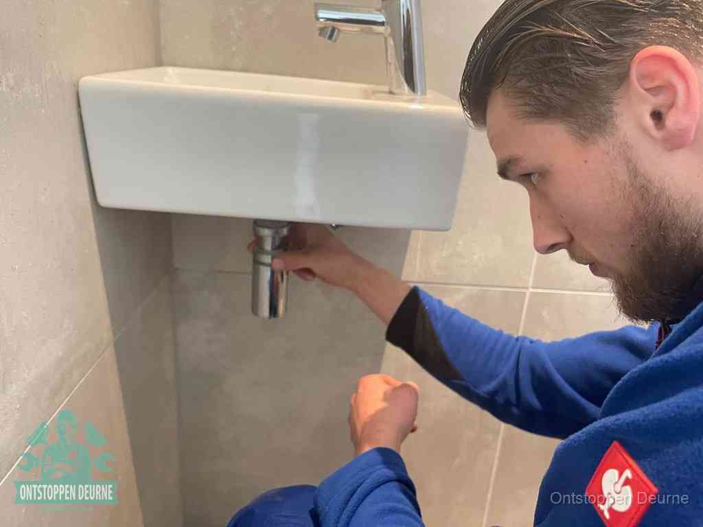 Een monteur draait met de hand de zwanenhalssifon onder een fonteinwasbak in de toiletruimte los om de bron van rioollucht in Deurne te vinden