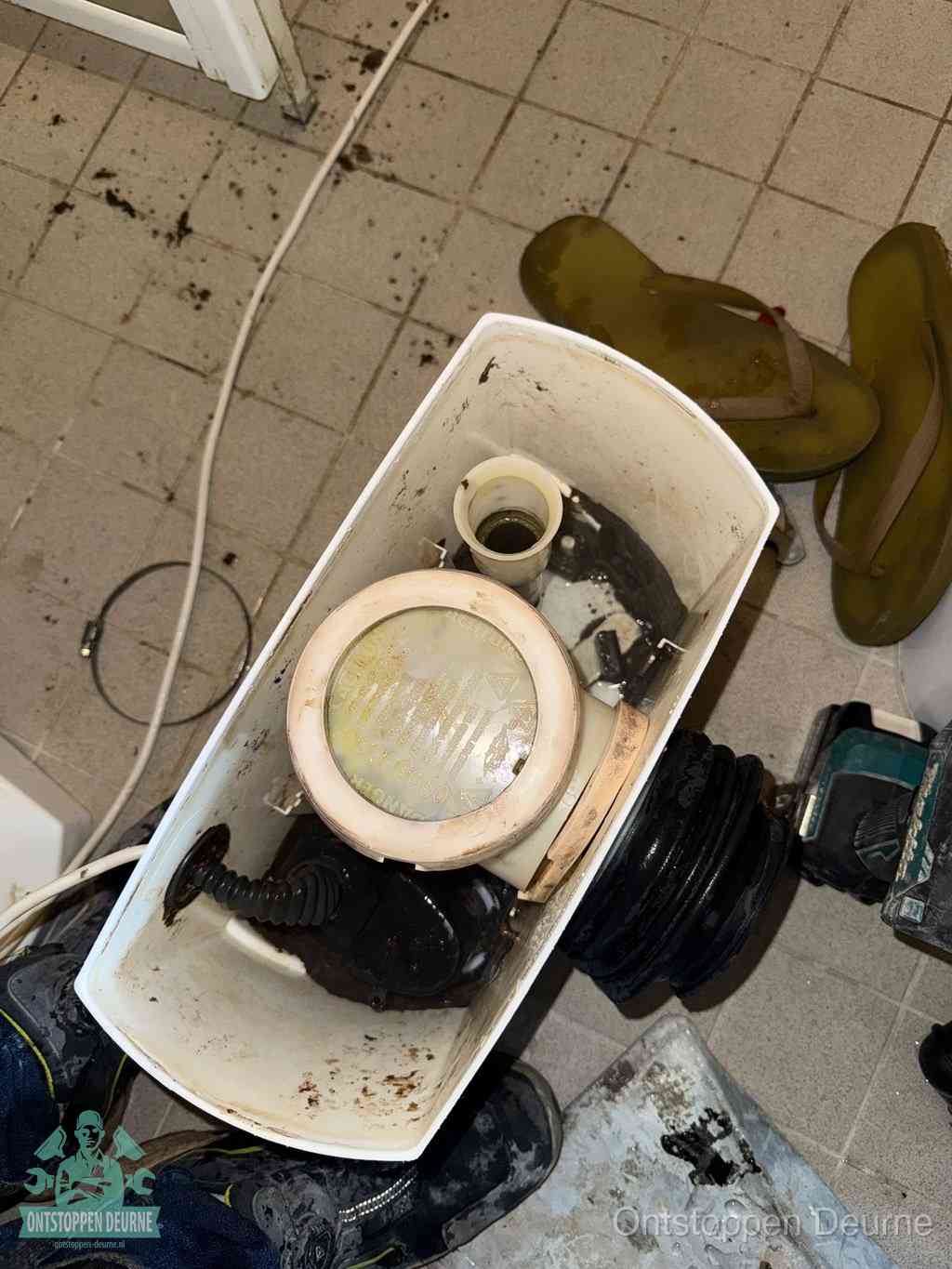 Een gedemonteerde toiletpot op de vloer met ontstoppingsveer en gereedschap ernaast, het verwijderen van een vastzittend voorwerp uit de wc in Deurne
