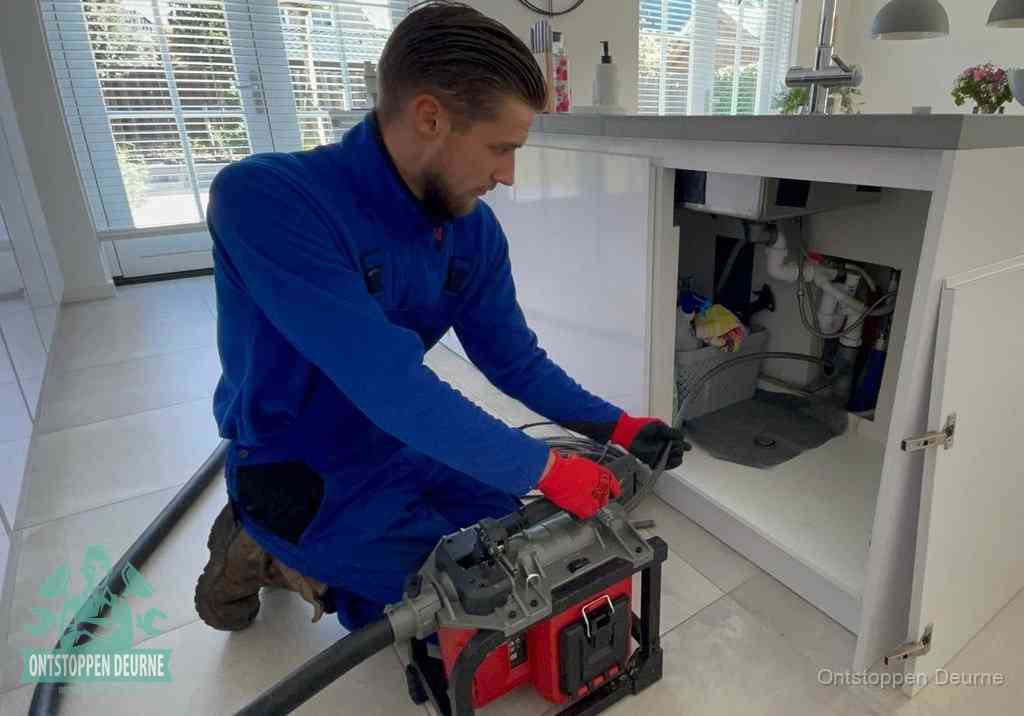 Een monteur voert een professionele elektrische ontstoppingsmachine in de afvoer onder de spoelbak tijdens het gootsteen ontstoppen in Deurne